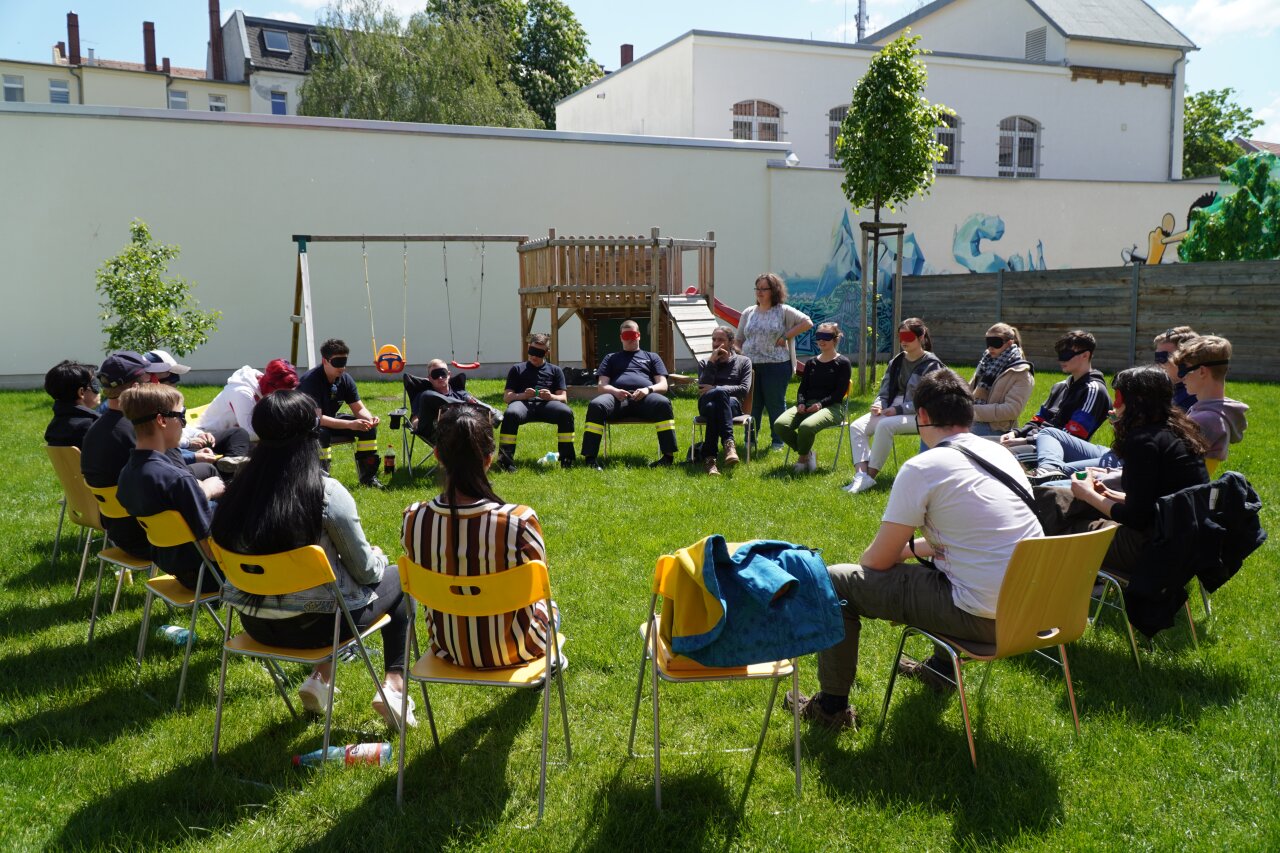 Viele Menschen mit verbundenen Augen sitzen auf Stühlen im Kreis.